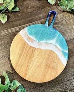Round wooden cutting board with a resin ocean wave design on one side, placed on a wooden surface with green leaves nearby.