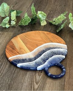 Round wooden serving board with blue and white resin art design, placed on a wooden surface with green leaves nearby.