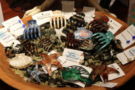 A wooden bowl holds assorted sea-themed hair clips, including shells, starfish, and claws, displayed on cards and stones.