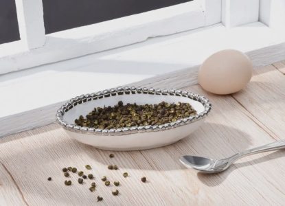 A bowl of green peppercorns, a whole egg, and a spoon on a wooden surface near a window.