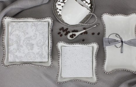 Rectangular white dishes with silver trim hold napkins and a ribbon, beside a white teacup, spoon, and coffee beans.