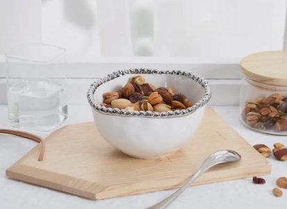 A white bowl of mixed nuts on a wooden board, next to a glass of water, a spoon, and a jar of more nuts.