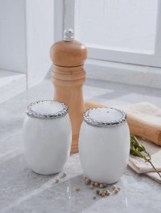 Two white ceramic salt and pepper shakers sit in front of a wooden pepper grinder on a kitchen counter.