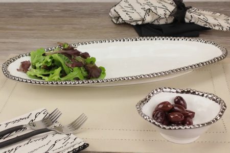 A white platter with mixed greens and a small bowl of olives on a placemat with cutlery and napkins nearby.