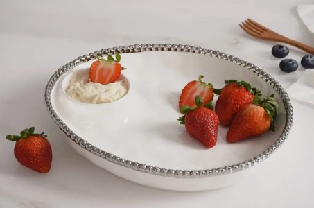A white platter with strawberries and a small section of cream, with blueberries and a wooden fork in the background.