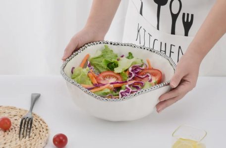 A person holding a bowl of fresh vegetable salad on a white table with a fork, tomato, and drink nearby.