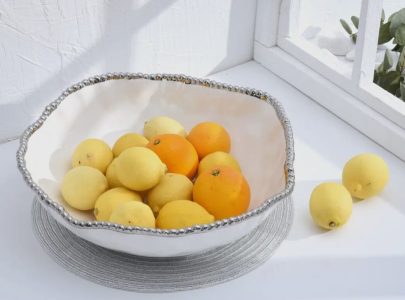 A white bowl with lemons and oranges sits on a gray placemat near a window; two lemons are outside the bowl.