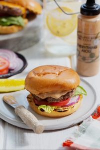 A cheeseburger with lettuce, tomato, and onion on a plate with a pickle spear, knife, and drink in the background.