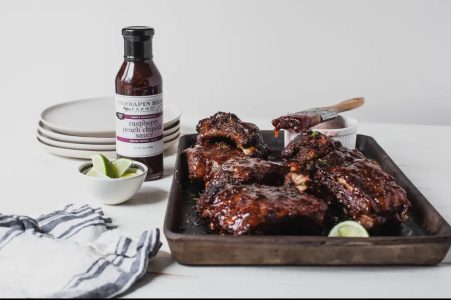 A tray of glazed ribs with lime wedges, a bottle of raspberry peach chipotle sauce, plates, and a striped napkin.