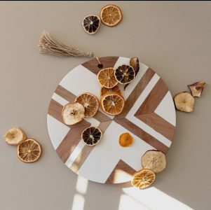 A round wooden and white decorative board with dried citrus slices arranged on and around it on a beige surface.