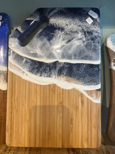 Rectangular wooden cutting board with a handle, featuring blue and white resin art resembling ocean waves on one end.