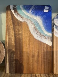 A wooden cutting board with a resin design resembling ocean waves and foam on one corner, displayed upright.