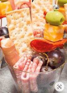 A clear plastic cup filled with crackers, cured meats, grapes, cheese cubes, and a green olive on a toothpick.