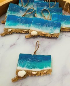 Resin ornaments shaped like Connecticut feature a beach scene with blue waves, sand, small shells, and twine loops for hanging.