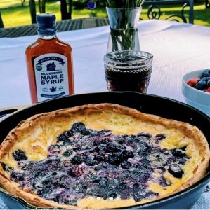 A baked blueberry pancake in a skillet with blueberry maple syrup, a glass of drink, and fresh berries on a table.