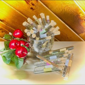 A glass bowl holds corked test tubes filled with herbs; three red apples with leaves rest beside more tubes on a light surface.