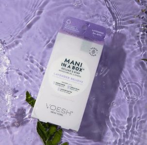 A box of VOESH Mani in a Box Lavender Relieve is placed on a lavender-colored surface with water and green leaves.