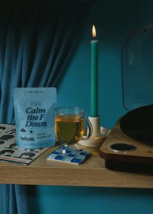 A glass mug of tea, a blue candle, and a packet labeled "Calm the F Down" sit on a wooden shelf with a record player.