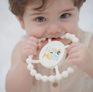 Baby with blue eyes holds and bites a round teething toy that says "I Love Mom" in colorful letters.