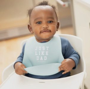 A baby wearing a blue bib that says "JUST LIKE DAD" sits in a high chair, looking at the camera.