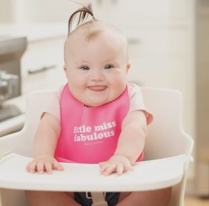 Baby with a ponytail wearing a pink bib that says "little miss fabulous," sitting in a high chair and smiling.