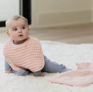 A baby in a pink bib and blue outfit sits on a white carpet next to a folded pink cloth in a bright room.