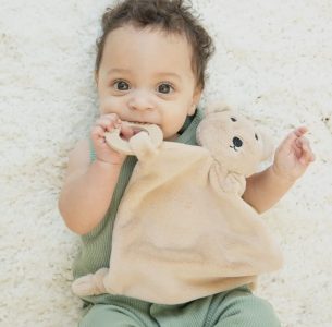 Baby lying on a soft, cream-colored rug, holding and chewing on a beige teddy bear blanket.